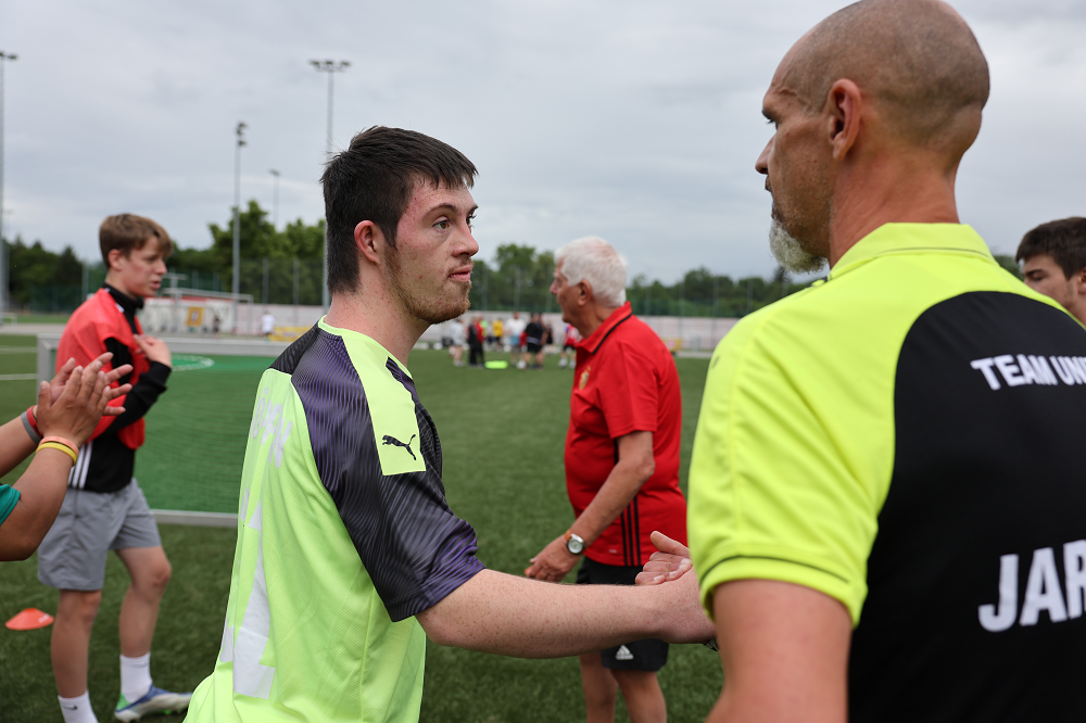 Zwei Männer, einer in grünem und einer in rotem T-Shirt, stehen auf einem Fußballfeld und unterhalten sich angeregt. Im Hintergrund sind weitere Personen und ein Fußballfeld zu sehen.