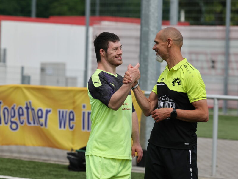 Zwei Männer in grünen Trikots geben sich auf dem Fußballplatz die Hände, im Hintergrund ein Banner mit der Aufschrift "Together we".