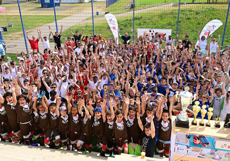 Eine große Gruppe von Kindern und Jugendlichen steht auf einem Fußballfeld und hebt die Hände in die Luft. Im Hintergrund sind weitere Personen und Pokale zu sehen. Es handelt sich um das Abschlussturnier des Projekts "Fußballtreff Kids" der DFL Stiftung.