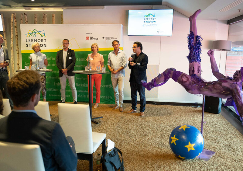 Ein Publikum sitzt vor einer Bühne, auf der mehrere Männer stehen und sprechen. Im Hintergrund ist ein Banner mit der Aufschrift "TeamEuropa" zu sehen. Rechts im Bild steht eine blaue Skulptur eines Akrobaten. Im Vordergrund sind helle Sessel und ein Tisch mit Dekoration zu sehen.