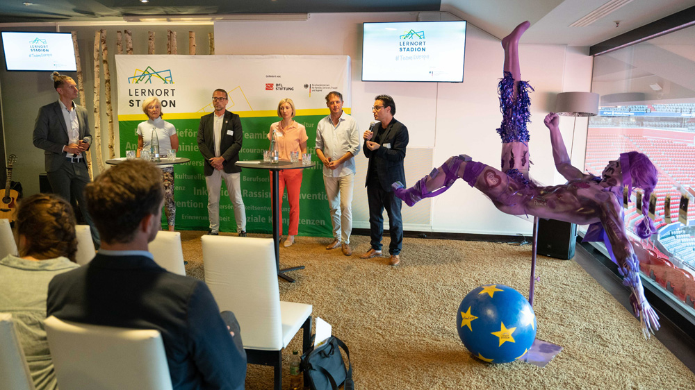 Ein Publikum sitzt vor einer Bühne, auf der mehrere Männer stehen und sprechen. Im Hintergrund ist ein Banner mit der Aufschrift "TeamEuropa" zu sehen. Rechts im Bild steht eine blaue Skulptur eines Akrobaten. Im Vordergrund sind helle Sessel und ein Tisch mit Dekoration zu sehen.