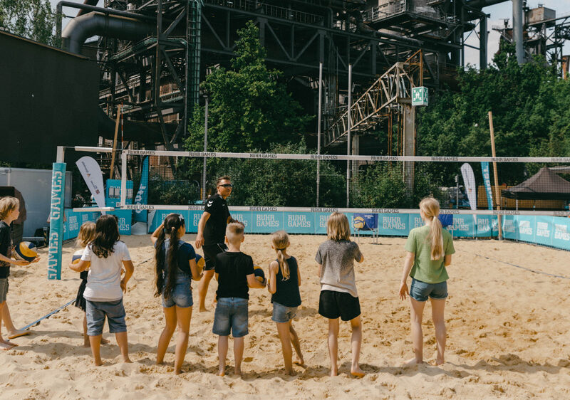 Eine Gruppe von Kindern und Jugendlichen steht auf einem Sandplatz und bildet einen Kreis. Im Hintergrund ist eine industrielle Kulisse mit einem alten Hochofen zu erkennen. Die Szene wirkt sonnig und positiv.