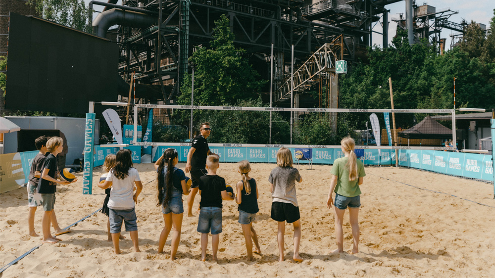 Eine Gruppe von Kindern und Jugendlichen steht auf einem Sandplatz und bildet einen Kreis. Im Hintergrund ist eine industrielle Kulisse mit einem alten Hochofen zu erkennen. Die Szene wirkt sonnig und positiv.
