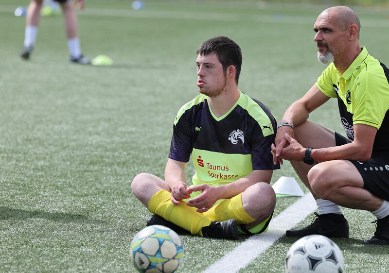 Zwei Männer sitzen auf dem Fußballrasen. Der jüngere Mann hockt mit verschränkten Beinen und blickt nachdenklich auf den Boden. Neben ihm sitzt ein älterer Mann in Hocke und beobachtet ihn. Im Hintergrund sind weitere Personen und Fußbälle zu sehen.