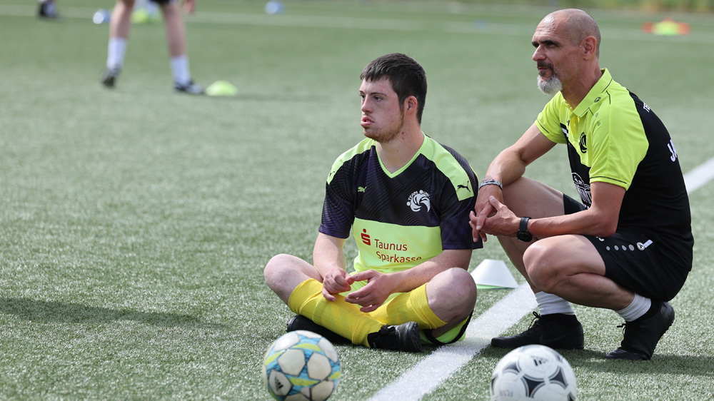 Zwei Männer sitzen auf dem Fußballrasen. Der jüngere Mann hockt mit verschränkten Beinen und blickt nachdenklich auf den Boden. Neben ihm sitzt ein älterer Mann in Hocke und beobachtet ihn. Im Hintergrund sind weitere Personen und Fußbälle zu sehen.