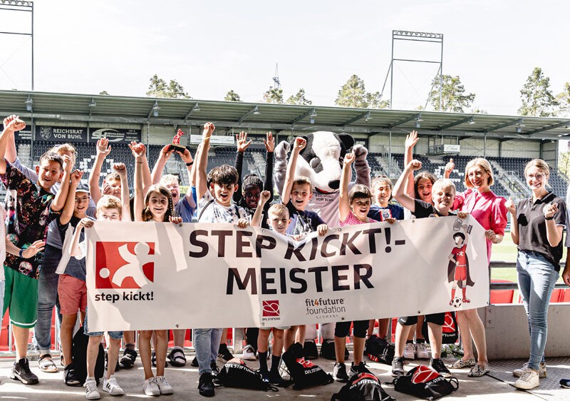 Eine Gruppe von Kindern und Jugendlichen steht strahlend mit hoch erhobenen Armen vor einer Tribüne. Sie halten ein Banner mit der Aufschrift "STEP KICKT! - MEISTER". Im Hintergrund ist ein Fußballstadion zu sehen.