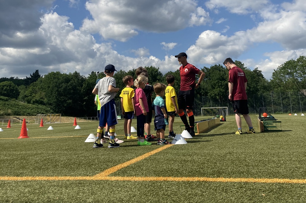 Gruppe von Kindern und Jugendlichen bei einer Fußball-Trainingseinheit auf einem grünen Rasenplatz, angeleitet von zwei Trainern. Im Hintergrund ein Wald.