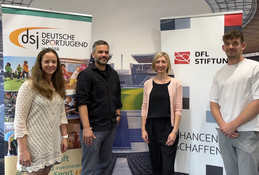 Vier Personen stehen nebeneinander vor einer Wand mit dem Logo der Deutschen Sportjugend und der DFL Stiftung. Von links nach rechts: Heike Hülse, Leon Ries, Franziska Fey und Lukas Bendix. Sie präsentieren das Rahmenkonzept „Kitas in Bewegung“.