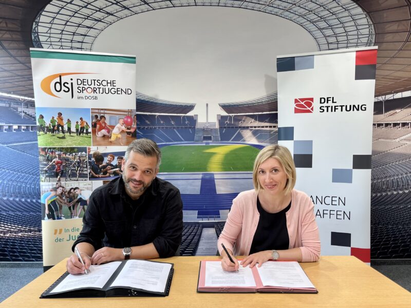 Leon Ries, Geschäftsführer der Deutschen Sportjugend, und Franziska Fey, Vorstandsvorsitzende der DFL Stiftung, unterzeichnen eine Vereinbarung an einem Tisch. Im Hintergrund sind Banner mit den Logos beider Organisationen und eine Stadionkulisse zu sehen.