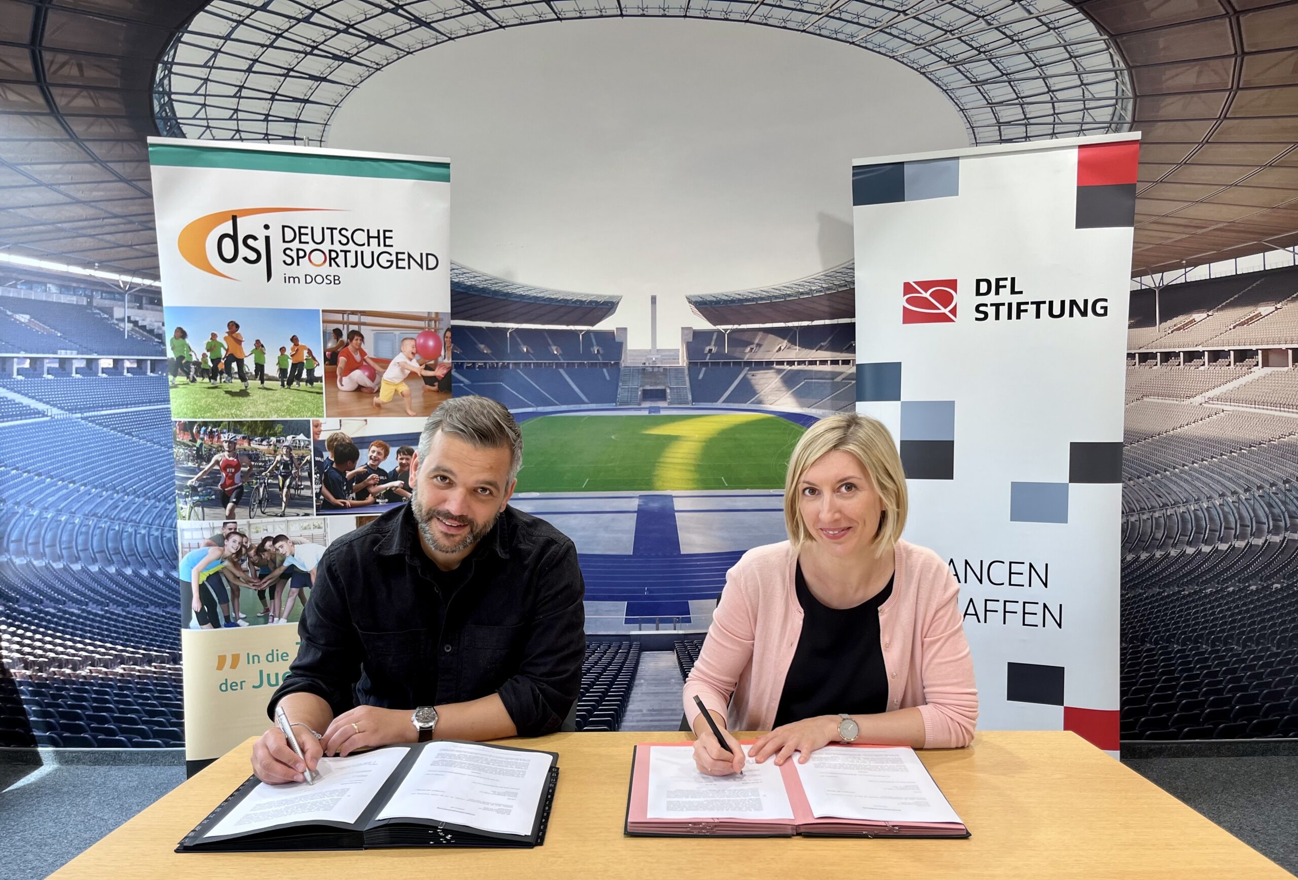 Leon Ries, Geschäftsführer der Deutschen Sportjugend, und Franziska Fey, Vorstandsvorsitzende der DFL Stiftung, unterzeichnen eine Vereinbarung an einem Tisch. Im Hintergrund sind Banner mit den Logos beider Organisationen und eine Stadionkulisse zu sehen.