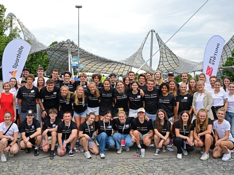 Eine große Gruppe junger Sportler*innen in schwarzer Sportbekleidung mit dem DFL-Stiftung-Logo posiert vor dem Olympiapark in München. Im Hintergrund ist das Dach der Olympiahalle zu sehen.