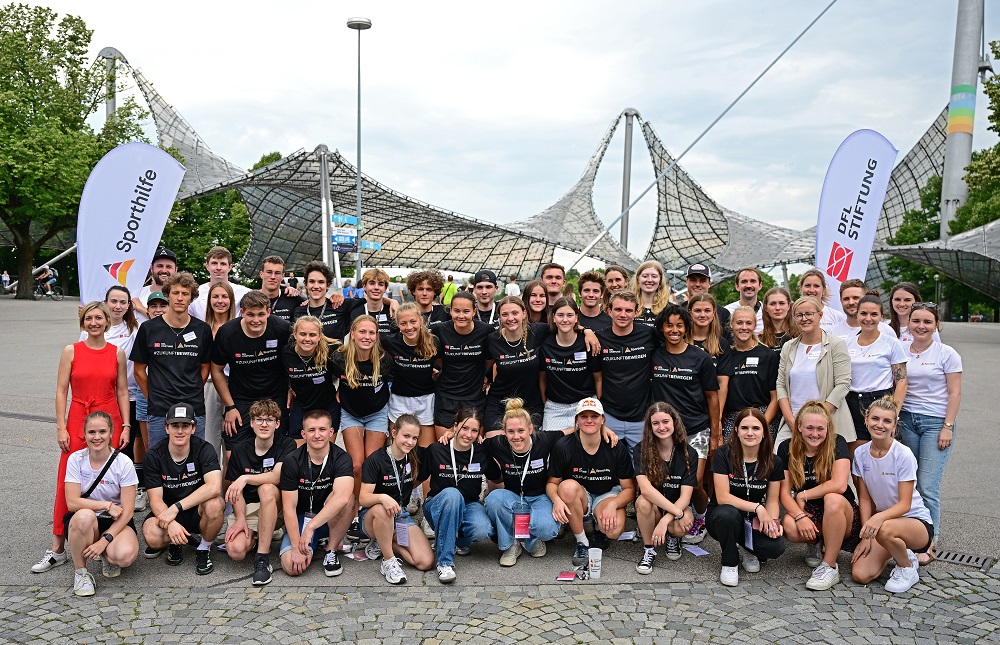 Eine große Gruppe junger Sportler*innen in schwarzer Sportbekleidung mit dem DFL-Stiftung-Logo posiert vor dem Olympiapark in München. Im Hintergrund ist das Dach der Olympiahalle zu sehen.