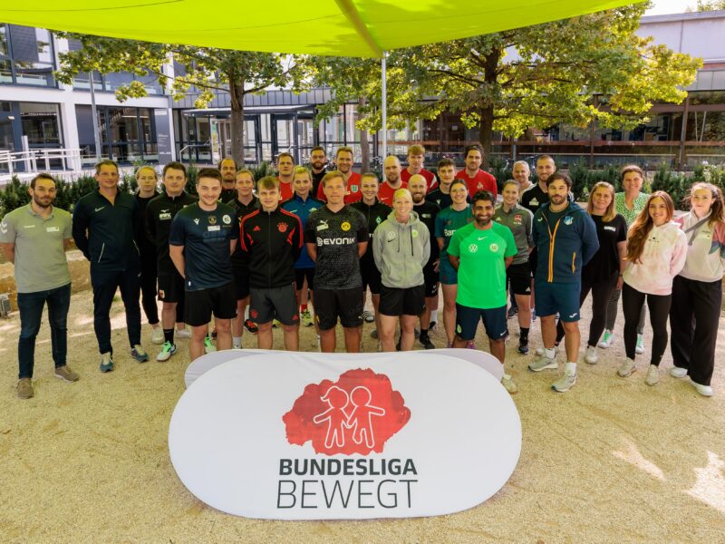Eine Gruppe von Teilnehmenden steht vor dem Institut für Sportwissenschaft der Universität Mainz zusammen. Im Vordergrund befindet sich ein Banner mit dem Logo "Bundesliga bewegt" und dem Symbol der DFL Stiftung. Die Teilnehmenden tragen Sportkleidung verschiedener Fußballvereine.