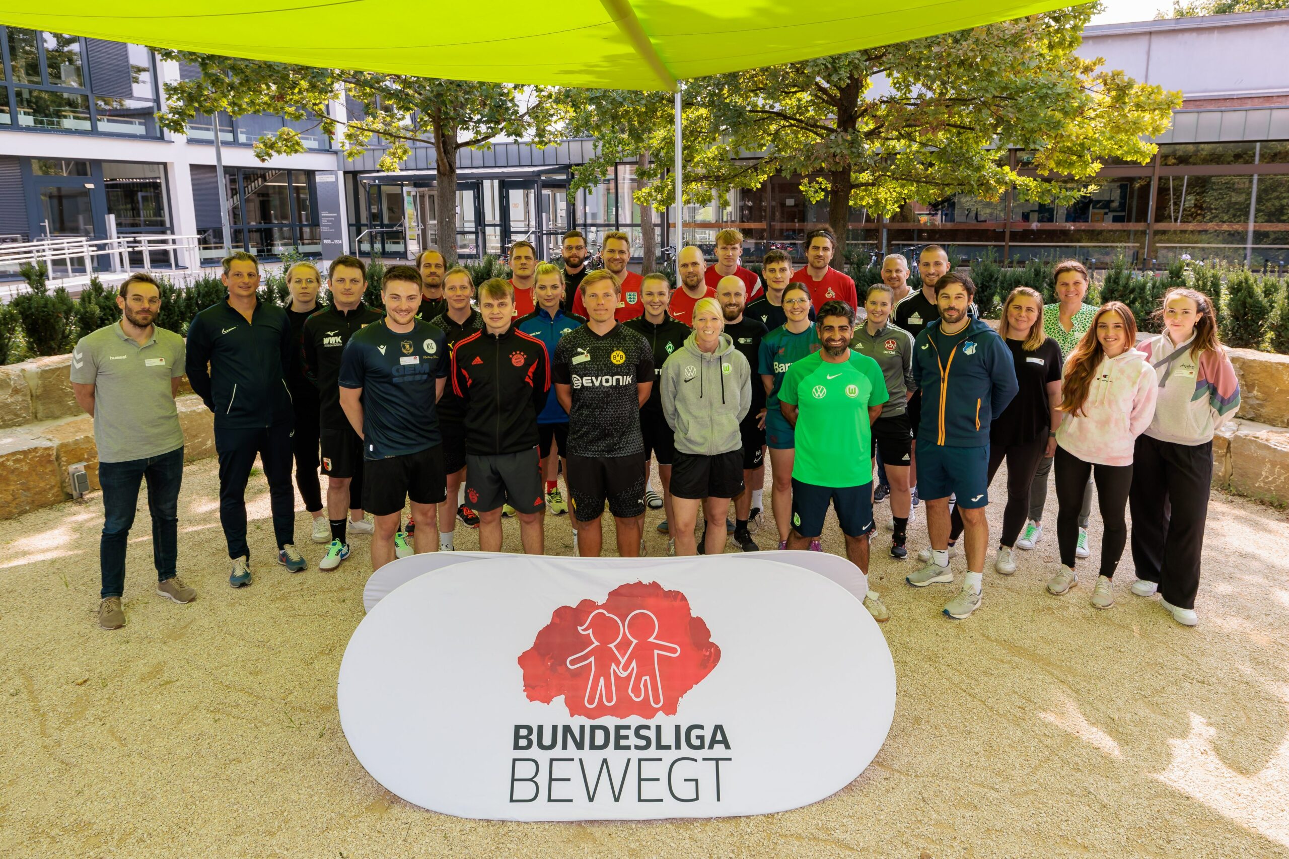 Eine Gruppe von Teilnehmenden steht vor dem Institut für Sportwissenschaft der Universität Mainz zusammen. Im Vordergrund befindet sich ein Banner mit dem Logo "Bundesliga bewegt" und dem Symbol der DFL Stiftung. Die Teilnehmenden tragen Sportkleidung verschiedener Fußballvereine.