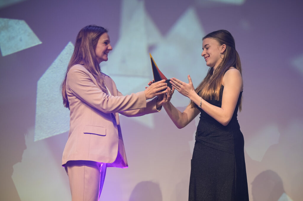 Tessa Lange nimmt eine Auszeichnung als gehörlose Leichtathletin des Jahres 2023 entgegen. Eine Frau überreicht ihr die Trophäe auf einer Bühne vor einem violett beleuchteten Hintergrund.