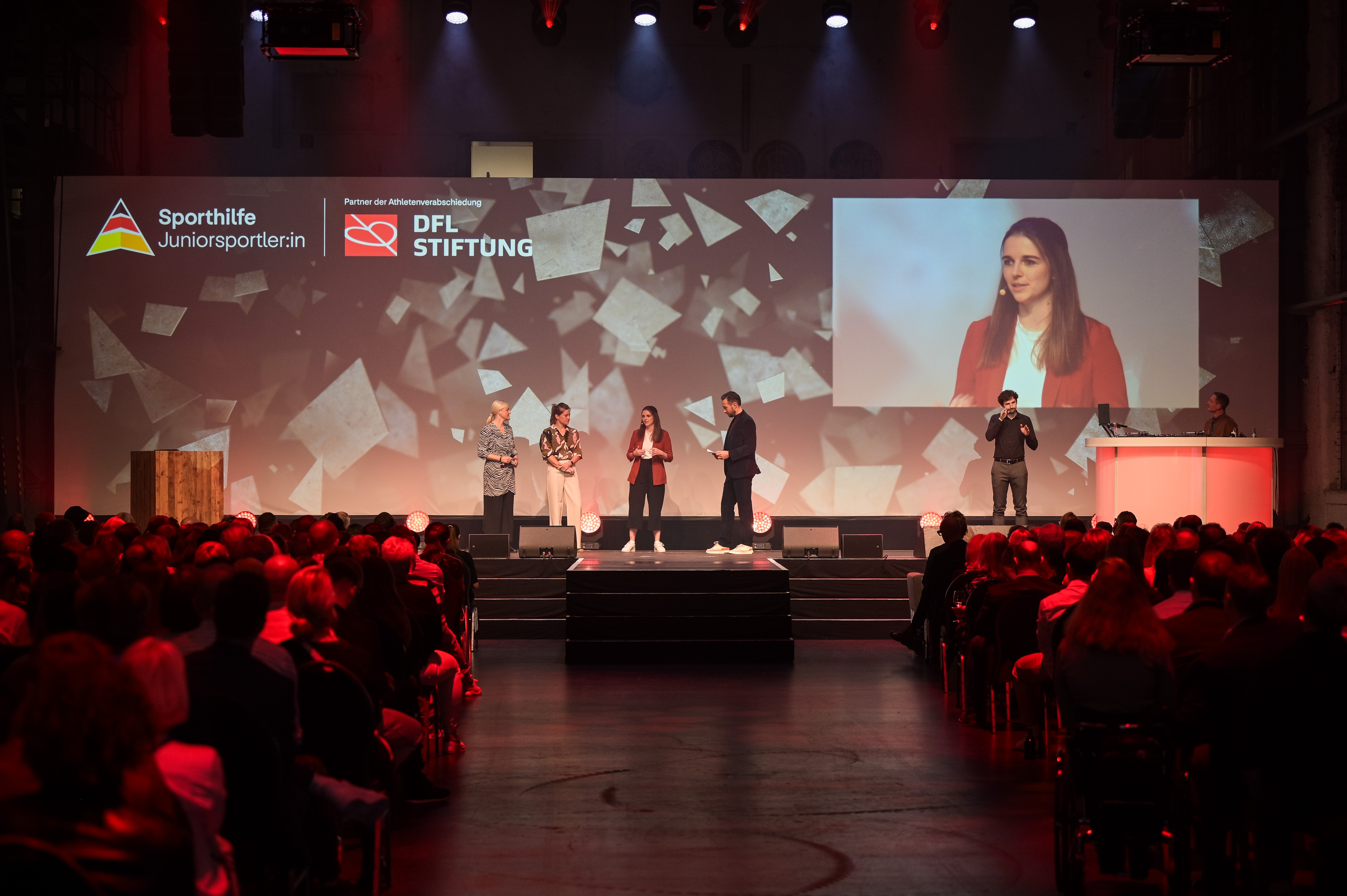 Ein Mann hält ein Mikrofon und interviewt eine Frau auf einer Bühne. Im Hintergrund ist auf einer großen Leinwand das Logo der Deutschen Sporthilfe und der DFL Stiftung zu sehen. Das Publikum ist in den unteren Bildbereich eingestreut.