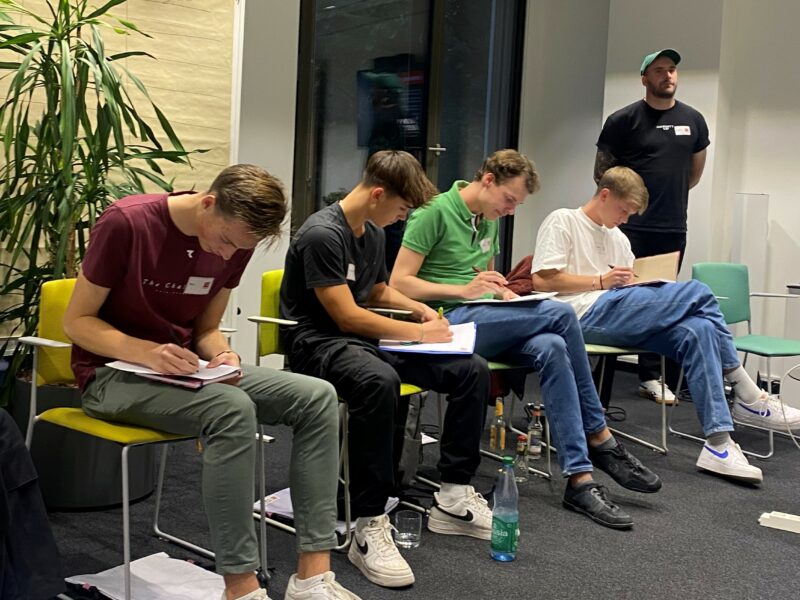 Eine Gruppe junger Menschen sitzt an Tischen und konzentriert sich auf Schreibtischmaterial, vermutlich im Rahmen eines Workshops oder einer Veranstaltung der DFL Stiftung.