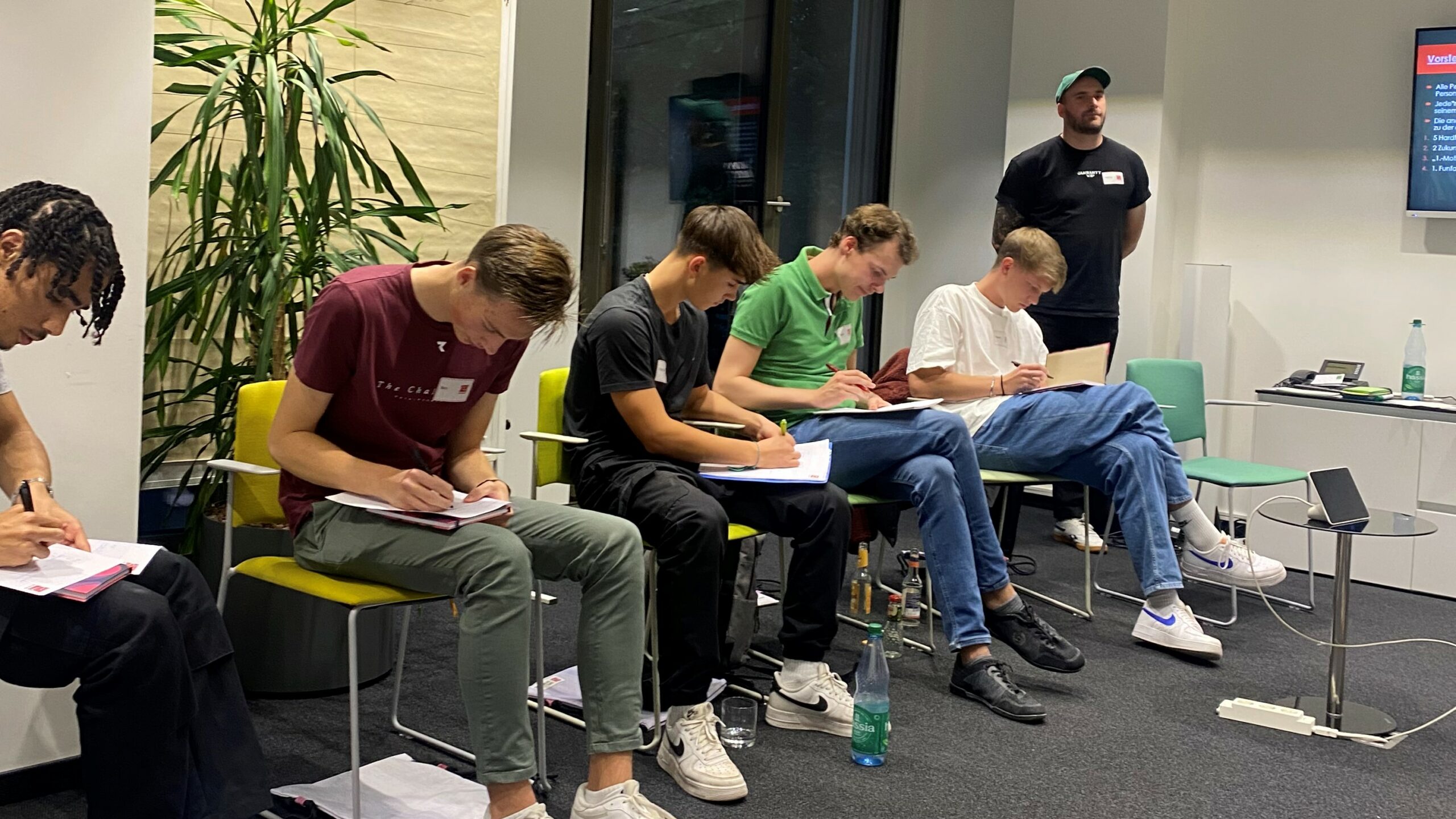 Eine Gruppe junger Menschen sitzt an Tischen und konzentriert sich auf Schreibtischmaterial, vermutlich im Rahmen eines Workshops oder einer Veranstaltung der DFL Stiftung.