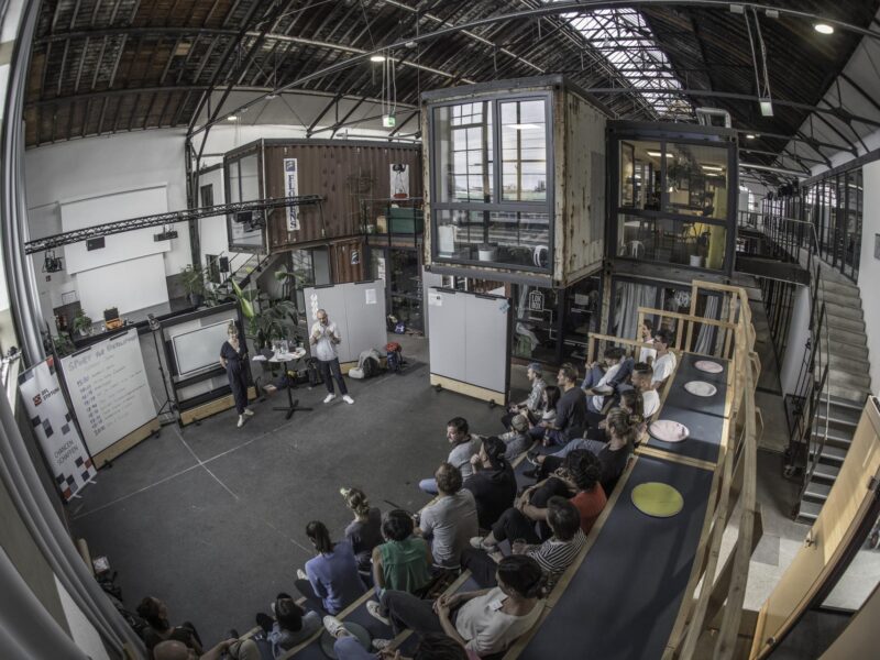 Ein weitwinkeliger Blick auf eine Veranstaltung der DFL Stiftung in einem modernen, offenen Raum in Freiburg. Eine Gruppe von Menschen sitzt auf Sitzkissen und schaut einem Redner zu, der vor einer Wand mit Flipchart und Präsentationsmaterial steht. Der Raum ist hell und industriell gestaltet mit sichtbaren Stahlträgern und Containern.