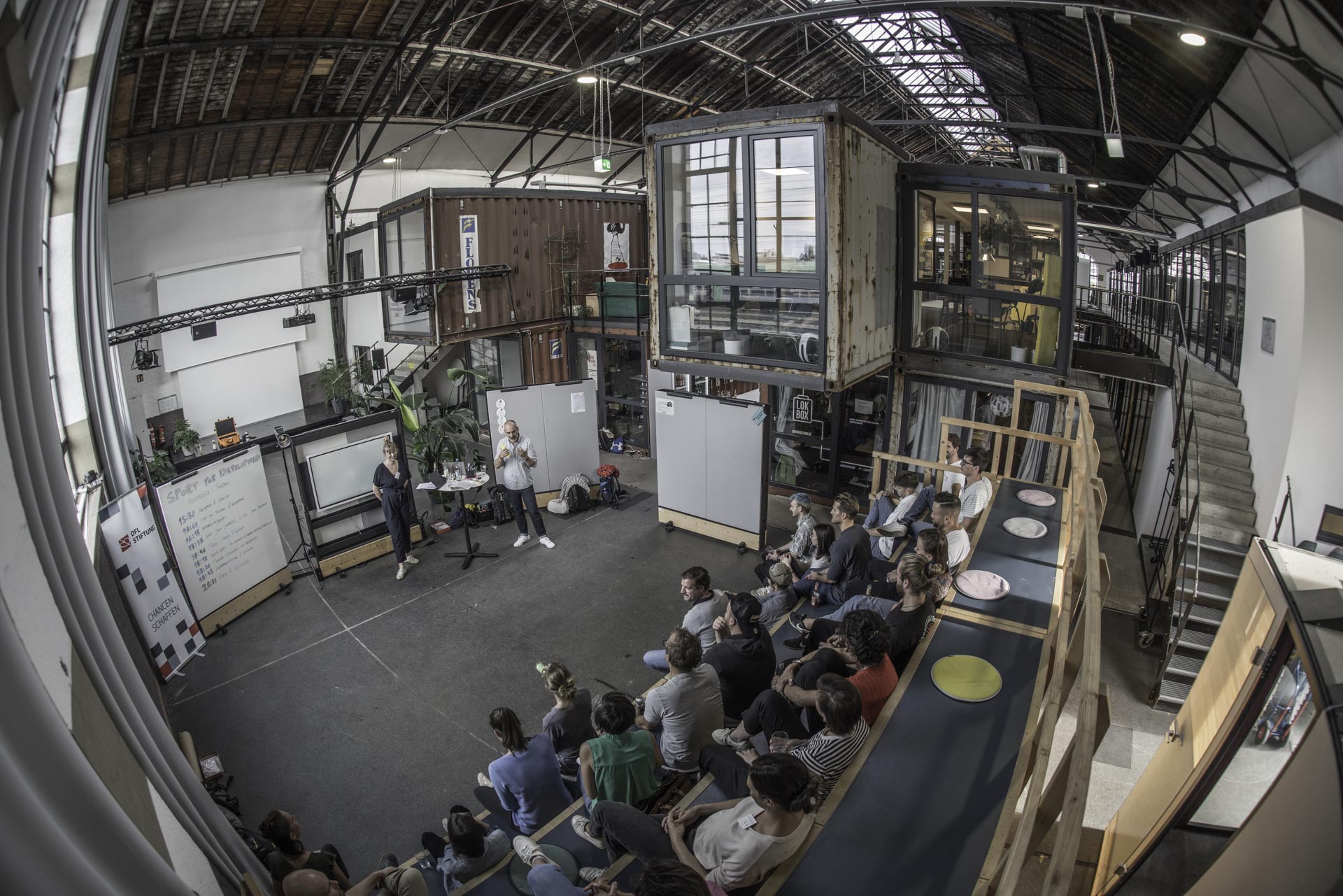 Ein weitwinkeliger Blick auf eine Veranstaltung der DFL Stiftung in einem modernen, offenen Raum in Freiburg. Eine Gruppe von Menschen sitzt auf Sitzkissen und schaut einem Redner zu, der vor einer Wand mit Flipchart und Präsentationsmaterial steht. Der Raum ist hell und industriell gestaltet mit sichtbaren Stahlträgern und Containern.