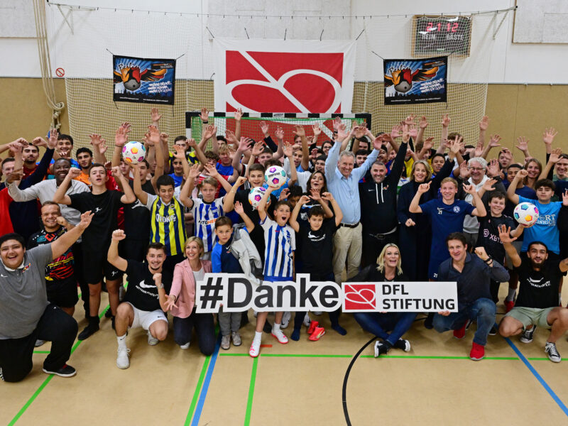 Eine große Gruppe von Menschen steht in einer Sporthalle zusammen und lächelt in die Kamera. Viele tragen sportliche Kleidung und halten Fußbälle. Im Hintergrund ist ein Banner mit der Aufschrift "#Danke DFL Stiftung" zu sehen. Das Bild zeigt einen Besuch des Kuratoriums der DFL Stiftung beim MitternachtsSport e.V. in Berlin am 13.10.2023.