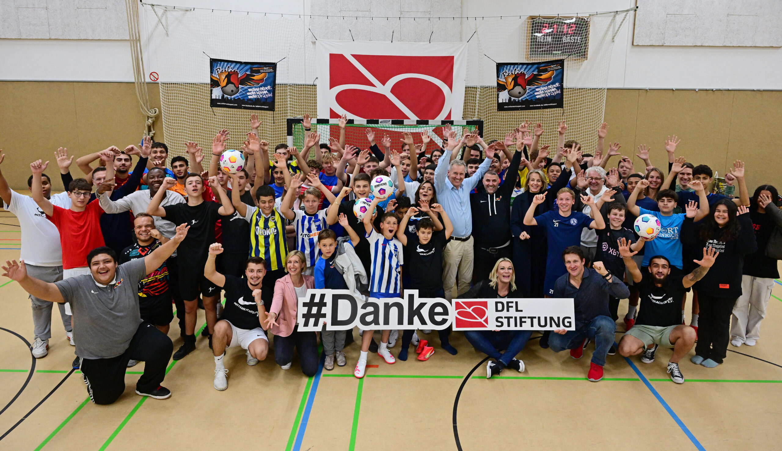 Eine große Gruppe von Menschen steht in einer Sporthalle zusammen und lächelt in die Kamera. Viele tragen sportliche Kleidung und halten Fußbälle. Im Hintergrund ist ein Banner mit der Aufschrift "#Danke DFL Stiftung" zu sehen. Das Bild zeigt einen Besuch des Kuratoriums der DFL Stiftung beim MitternachtsSport e.V. in Berlin am 13.10.2023.