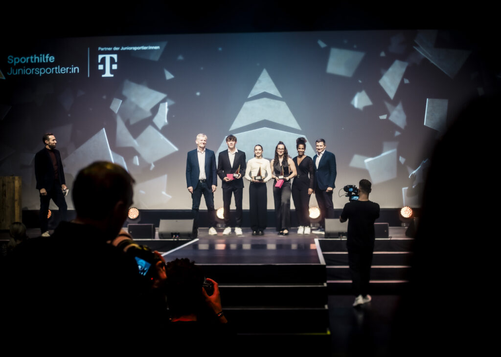 Preisträger*innen der Deutschen Sporthilfe "Juniorsportler*in des Jahres 2023" stehen auf einer Bühne und halten ihre Auszeichnungen entgegen. Im Hintergrund ist ein abstraktes Lichtdesign zu sehen.