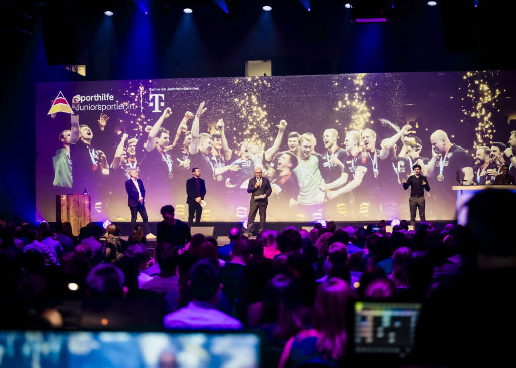 U21-Handball-Junioren stehen auf einer Bühne und werden für ihre Leistung als Mannschaft des Jahres geehrt. Im Hintergrund eine Leinwand mit dem Logo der Deutschen Sporthilfe und des Sponsors T-Mobile.