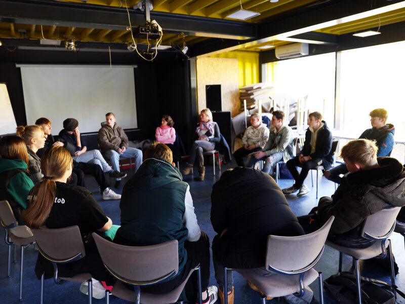 Gruppe junger Menschen sitzt im Kreis auf Stühlen in einem hellen Raum und diskutiert. Im Hintergrund eine Leinwand und ein Fenster mit Tageslicht.