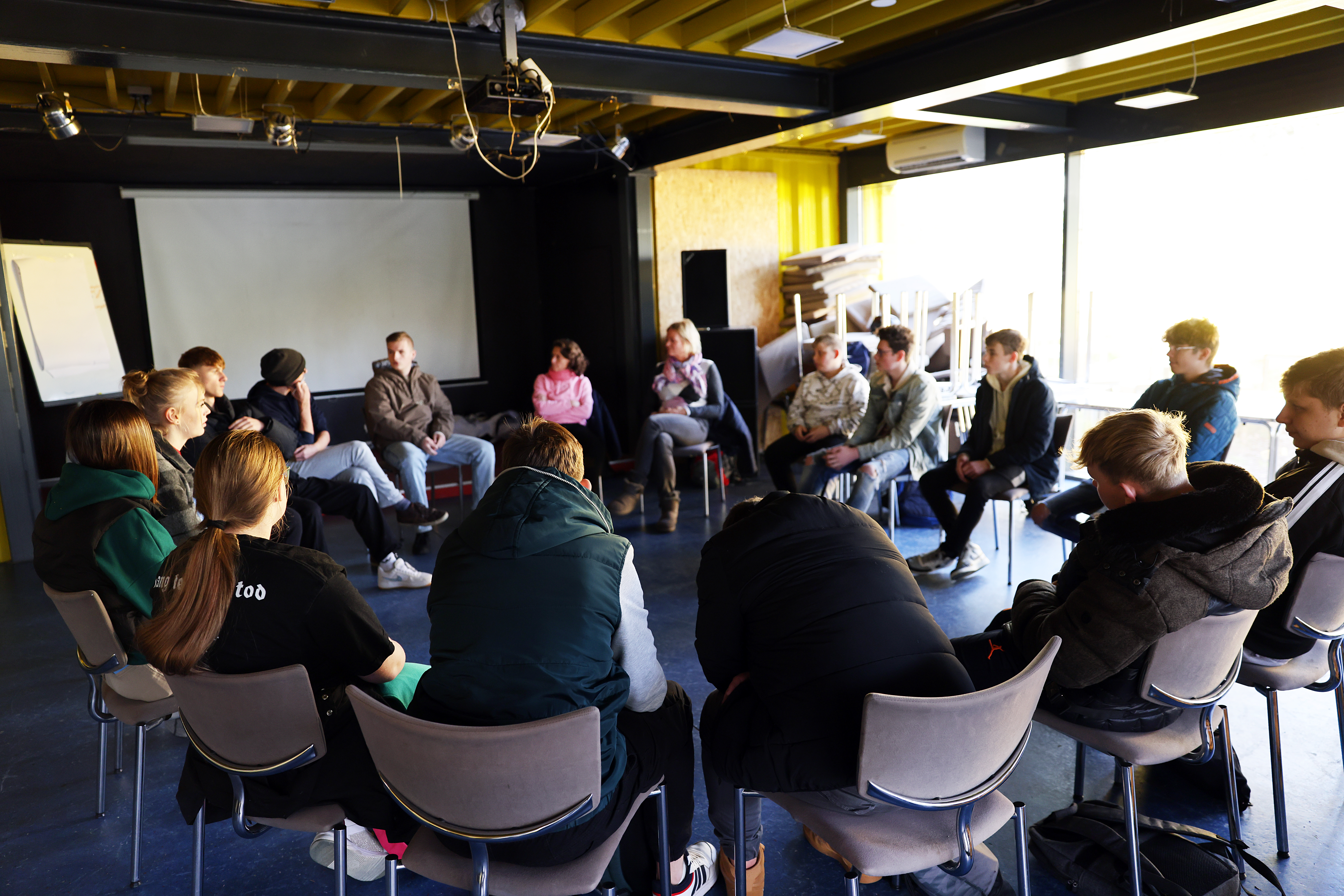 Gruppe junger Menschen sitzt im Kreis auf Stühlen in einem hellen Raum und diskutiert. Im Hintergrund eine Leinwand und ein Fenster mit Tageslicht.