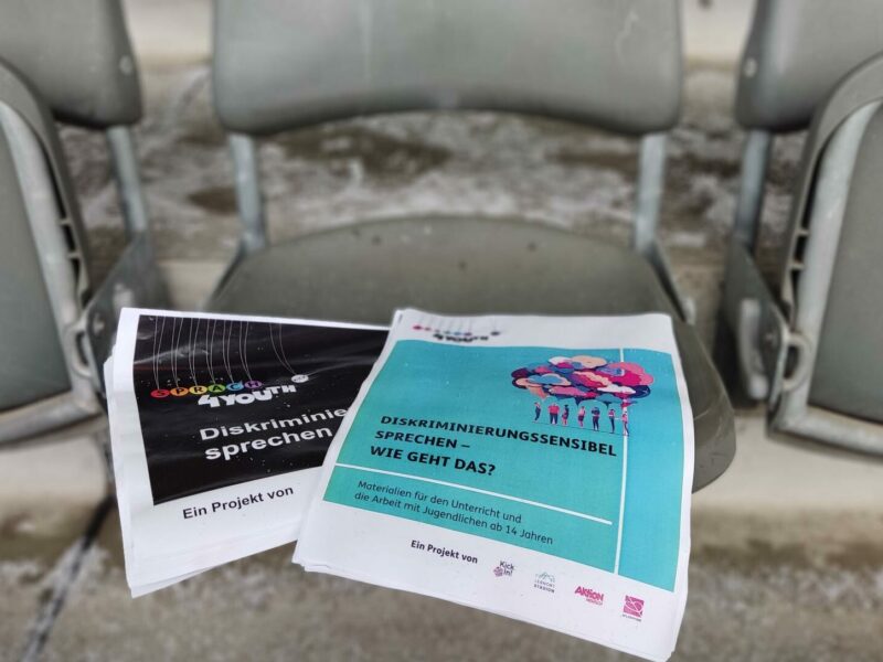 Zwei Flyer liegen auf einem grauen Sessel im Olympiastadion. Der eine Flyer ist schwarz mit dem Aufdruck "Diskriminierung sprechen", der andere blau mit dem Titel "Diskriminierungssensibel – Wie geht das?". Darunter steht "Materialien für die Arbeit mit Jugendlichen ab 14 Jahren". Beide Flyer tragen den Hinweis "Ein Projekt von Kick".