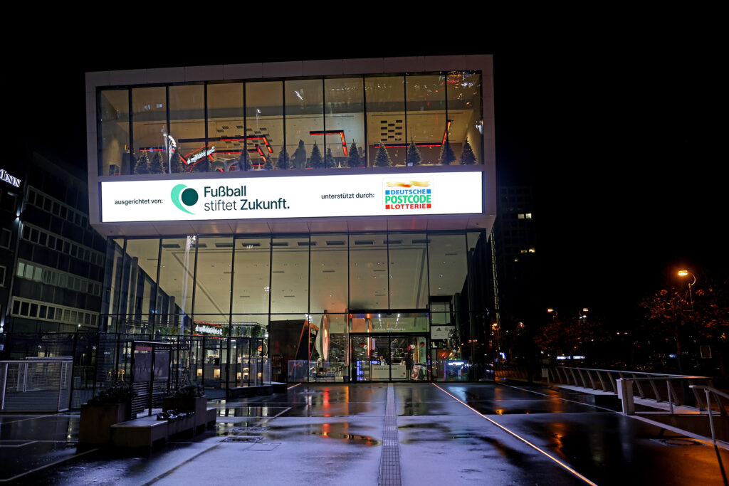 Nachtsaufnahme eines modernen Gebäudes mit einer großen LED-Leinwand. Auf der Leinwand steht in großen Buchstaben "Fußball stiftet Zukunft" und darunter das Logo der Deutsche Postcode Lotterie. Vor dem Gebäude befindet sich ein beleuchteter Platz mit Bäumen und Personen.