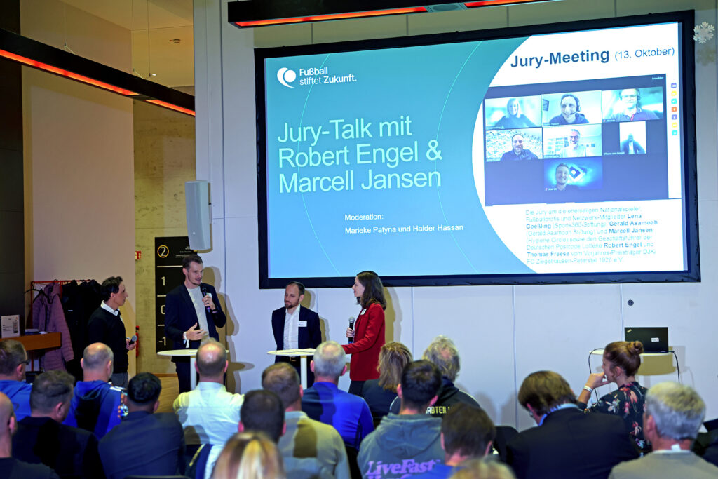 Ein Saal gefüllt mit Zuschauern, während auf einer großen Leinwand "Jury-Talk mit Robert Engel & Marcell Jansen" steht. Robert Engel und Marcell Jansen stehen auf einer Bühne und führen ein Gespräch, während im Hintergrund eine Collage mit Porträts von Jurymitgliedern zu sehen ist.