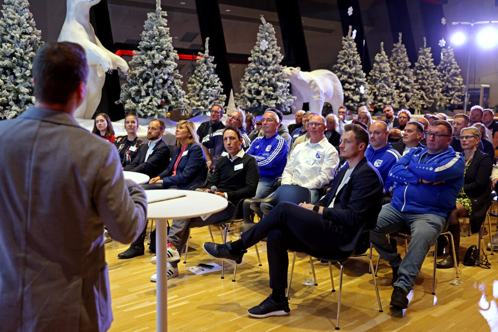 Zuhörer sitzen auf Stühlen in einem hellen Raum, während eine Person mit dem Rücken zum Betrachter zu sehen ist und spricht. Im Hintergrund sind verschneite Tannenbäume und Eisbärenfiguren zu erkennen.
