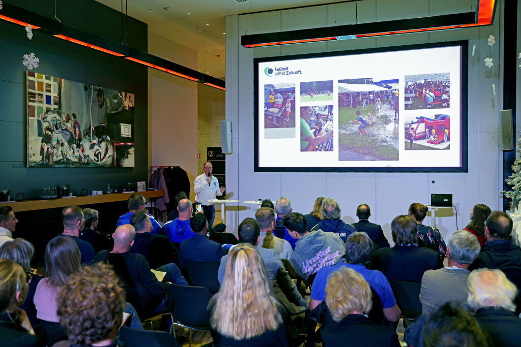 Ein Mann steht vor einer großen Leinwand und hält einen Vortrag vor einem Publikum. Auf der Leinwand sind verschiedene Bilder zu sehen, die offenbar Projekte der DFL Stiftung illustrieren. Im Hintergrund sind Kunstwerke an der Wand zu erkennen.