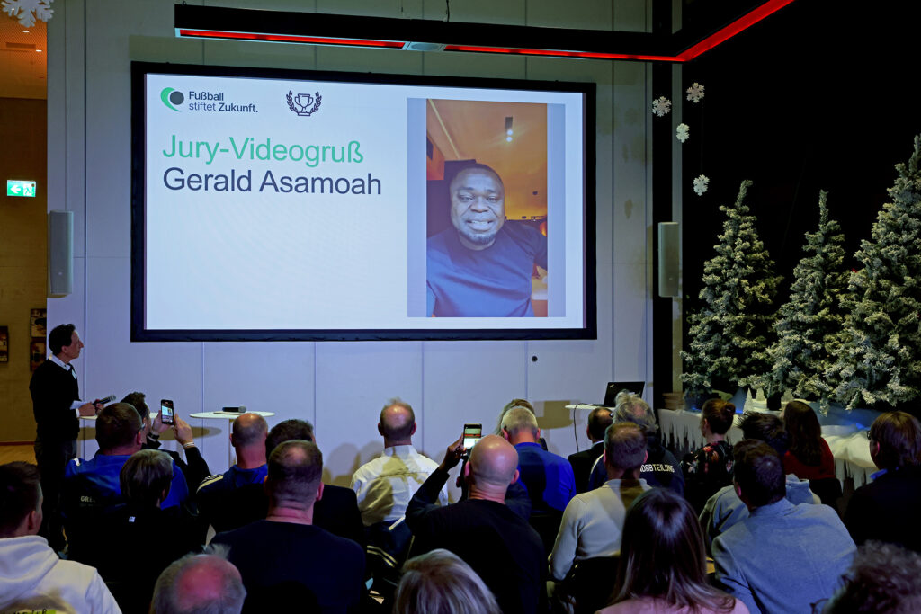 Bildschirm mit Porträt von Gerald Asamoah während einer Veranstaltung der DFL Stiftung, im Publikum sind Personen mit Smartphones zu sehen.
