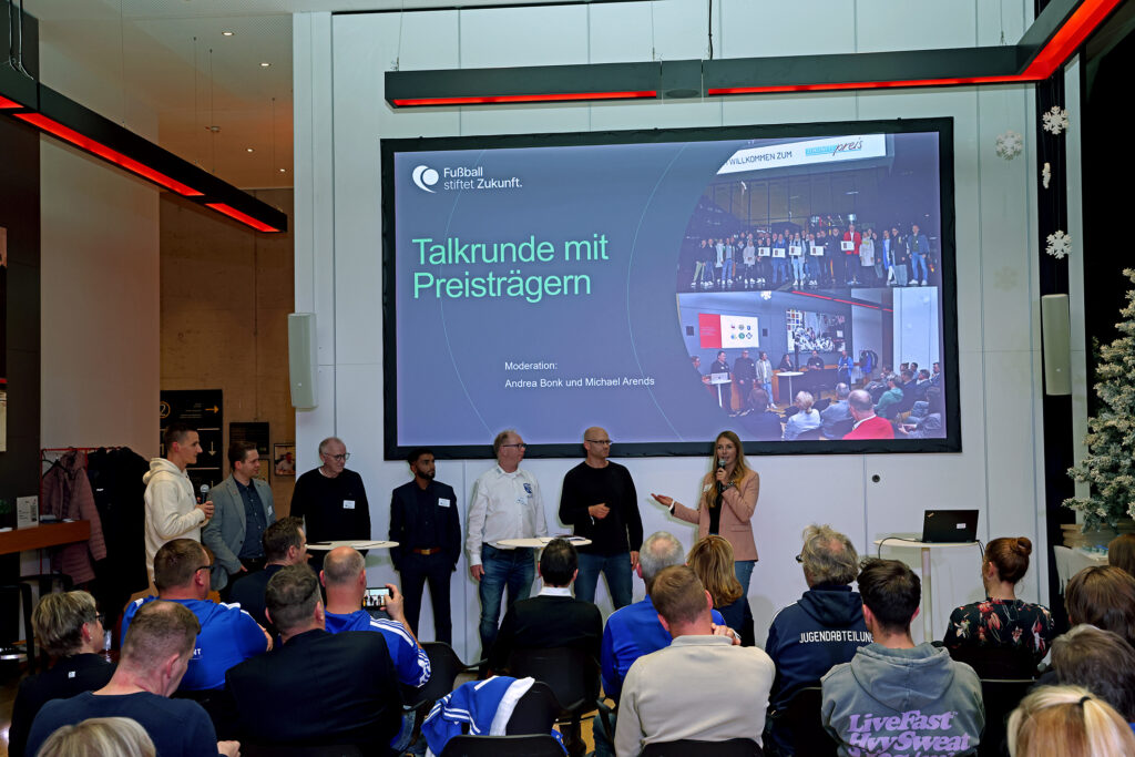 Eine Frau steht auf einer Bühne vor einer großen Leinwand und hält eine Rede. Auf der Leinwand steht "Fußball stiftet Zukunft. Talkrunde mit Preisträgern" sowie die Namen der Moderatoren Andrea Berk und Michael Arends. Im Publikum sitzen und stehen zahlreiche Personen, darunter einige in Sportkleidung mit Aufdruck "LiveFast". Die Szene wirkt wie eine Preisverleihung oder eine Podiumsdiskussion der DFL Stiftung.