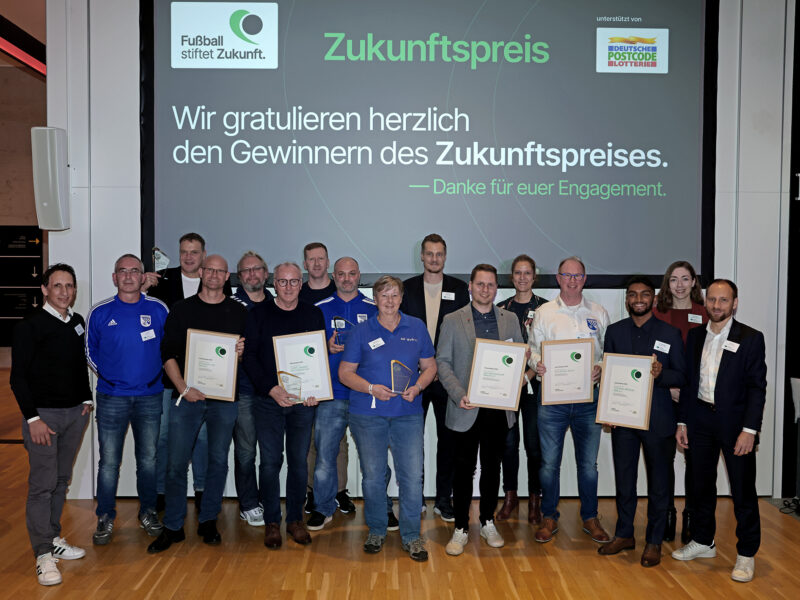 Eine Gruppe von etwa 15 Personen steht auf einer Bühne und hält Urkunden in den Händen. Im Hintergrund ein Banner mit der Aufschrift "Zukunftspreis" und dem Logo der DFL Stiftung sowie dem Sponsor "Deutsche Postcode Lotterie". Es handelt sich um die Preisträger*innen des Zukunftspreises 2023.