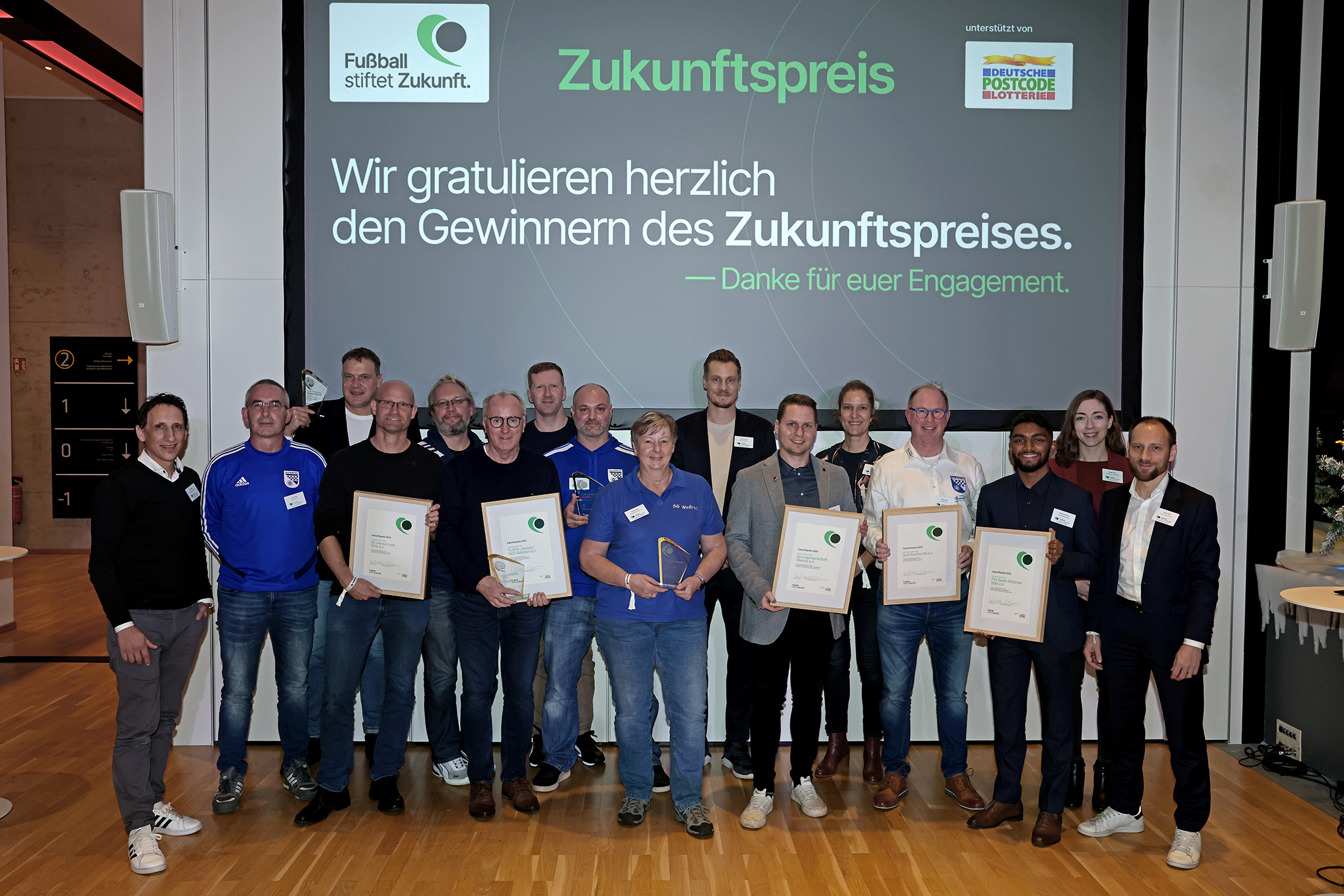 Eine Gruppe von etwa 15 Personen steht auf einer Bühne und hält Urkunden in den Händen. Im Hintergrund ein Banner mit der Aufschrift "Zukunftspreis" und dem Logo der DFL Stiftung sowie dem Sponsor "Deutsche Postcode Lotterie". Es handelt sich um die Preisträger*innen des Zukunftspreises 2023.