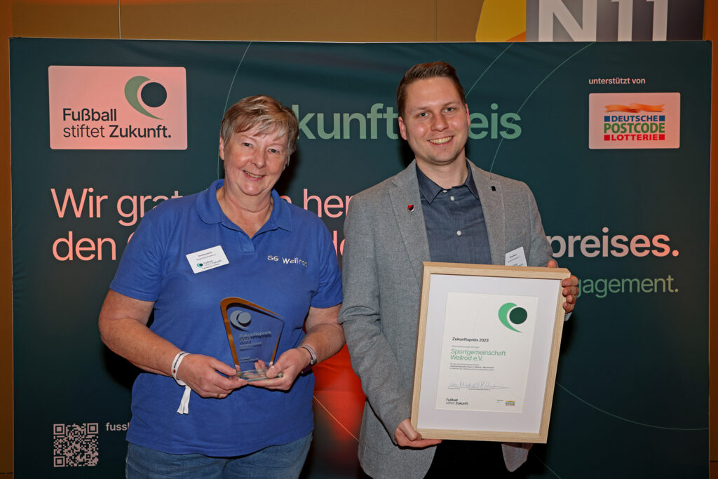 Eine Frau und ein Mann halten jeweils eine gläserne Trophäe und eine Urkunde mit dem Aufdruck "ZukunftsPreis 2023" in den Händen. Im Hintergrund ist ein Banner mit der Aufschrift "Fußball stiftet Zukunft" und dem Logo der Deutschen Postcode Lotterie zu sehen.