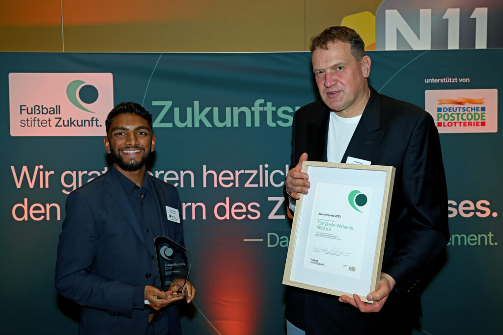 Zwei Männer stehen lächelnd mit einem Preis in der Hand vor einer Wand mit dem Schriftzug "Zukunftspreis". Der linke Mann hat dunkle Haare und einen Bart, der rechte Mann ist älter und hat graues Haar. In ihren Händen halten sie eine Urkunde mit dem Aufdruck "Zukunftspreis" und dem Logo der DFL Stiftung sowie der Deutschen Postcode Lotterie.