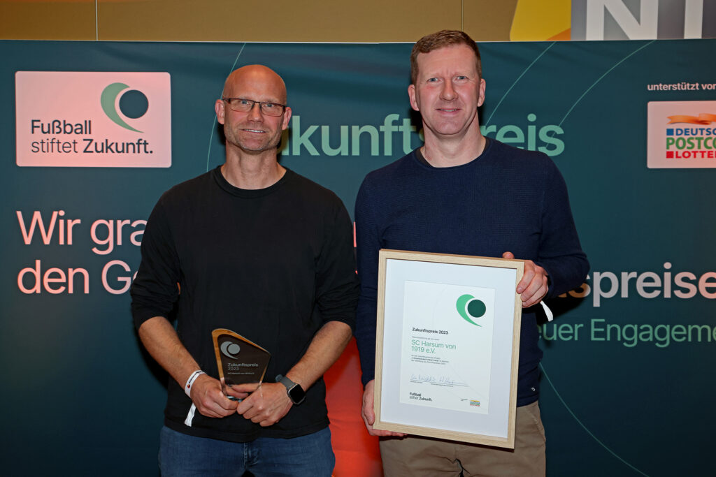 Zwei Männer halten jeweils eine Auszeichnung des "Zukunftspreises" in den Händen, im Hintergrund ein Banner mit der Aufschrift "Fußball stiftet Zukunft".
