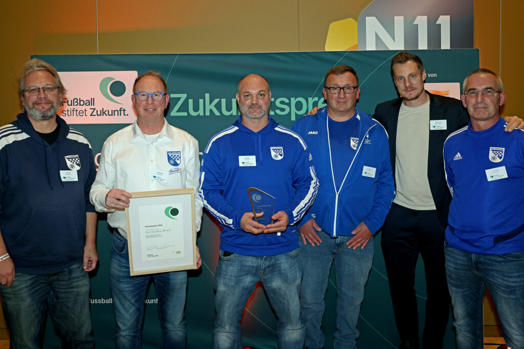 Eine Gruppe von Männern steht lächelnd in einer Turnhalle zusammen und hält eine Auszeichnung entgegen. Im Hintergrund ist ein Banner mit der Aufschrift "Zukunft" und dem Logo der DFL Stiftung zu sehen.