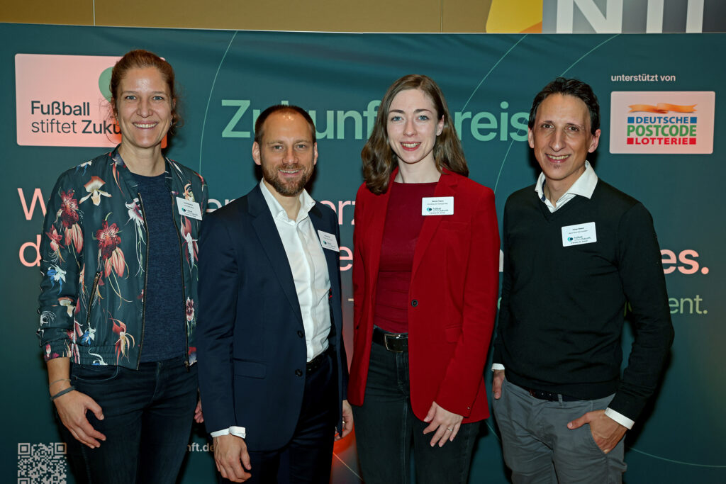 Vier Personen stehen vor einem Hintergrund mit der Aufschrift "Fußball stiftet Zukunft". Von links nach rechts: Eine Frau in einem gemusterten Oberteil, ein Mann im blauen Anzug, eine Frau in einem roten Kleid und ein Mann im schwarzen Anzug. Alle tragen Namensschilder. Im Hintergrund ist das Logo der Deutschen Postcode Lotterie zu sehen.