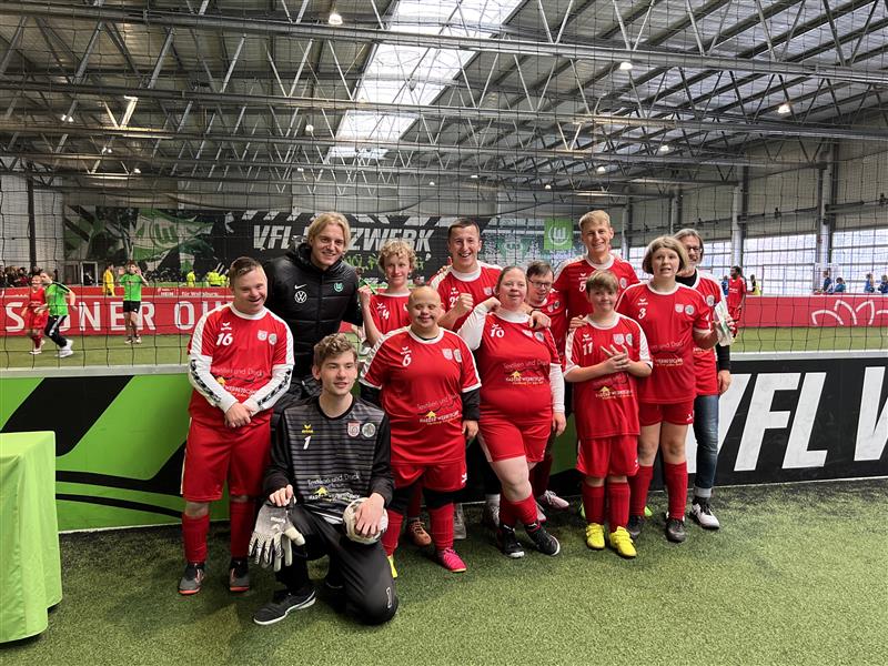 Eine Gruppe von Fußballspielerinnen und ein Betreuer posieren in einer Sporthalle. Sie tragen rote Trikots mit weißen Details und schwarze Shorts. Im Hintergrund sind Werbebanner und die Hallenstruktur zu sehen.