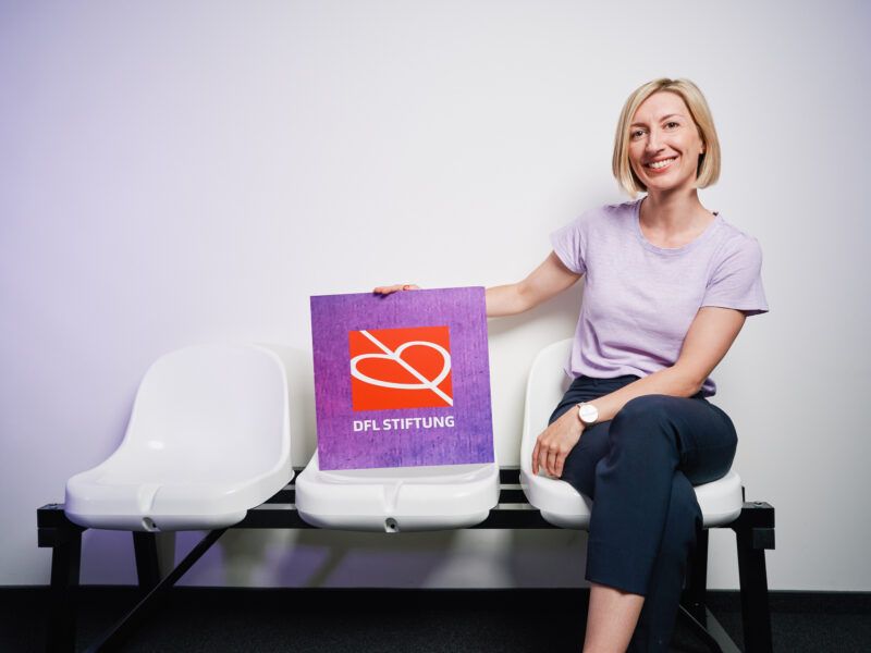 Franziska Fey sitzt lächelnd auf einer weißen Bank vor einer hellen Wand und hält ein violettes Plakat mit dem Logo der DFL Stiftung.