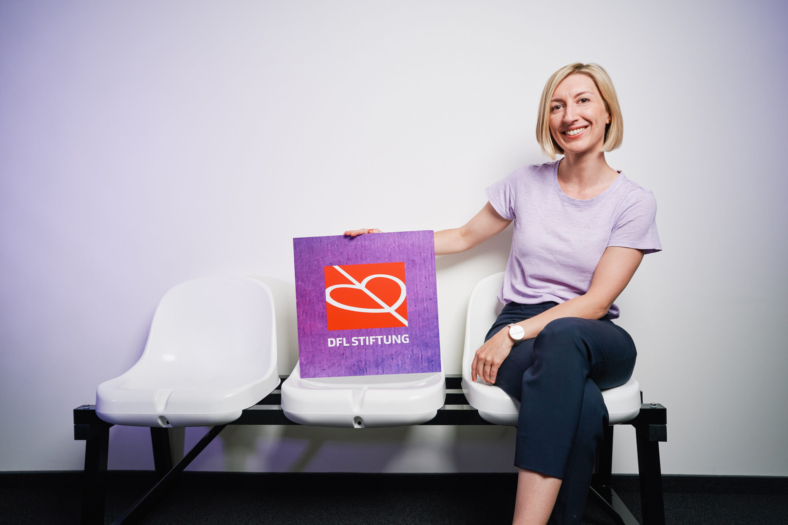 Franziska Fey sitzt lächelnd auf einer weißen Bank vor einer hellen Wand und hält ein violettes Plakat mit dem Logo der DFL Stiftung.