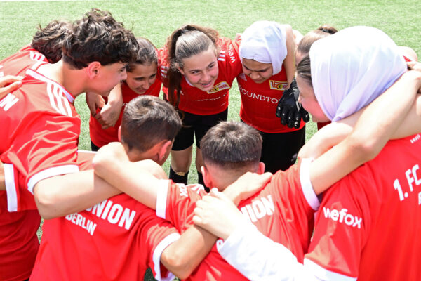 Eine Gruppe von Jugendlichen, gemischt geschlechtlich und mit unterschiedlichem kulturellem Hintergrund, steht im Kreis auf einem Fußballplatz und umarmt sich. Sie tragen rote T-Shirts mit dem Logo "1. FC Union Berlin" und "WeForx". Im Hintergrund ist grünes Gras zu sehen.