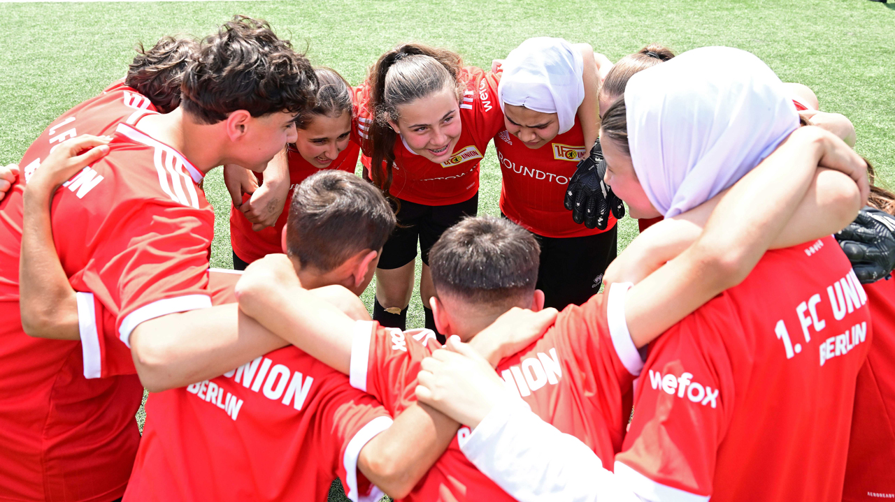 Eine Gruppe von Jugendlichen, gemischt geschlechtlich und mit unterschiedlichem kulturellem Hintergrund, steht im Kreis auf einem Fußballplatz und umarmt sich. Sie tragen rote T-Shirts mit dem Logo "1. FC Union Berlin" und "WeForx". Im Hintergrund ist grünes Gras zu sehen.