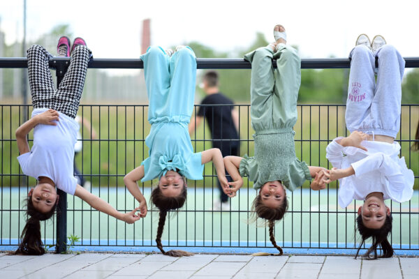 Vier Mädchen hängen an einer Stange und machen den Handstand, im Hintergrund eine Sportfläche und verschwommene Personen.